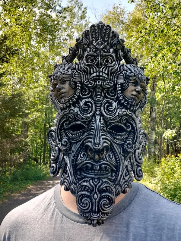Osoba s detailnou ornamentálnou maskou stojí v lese, vytvára tajomnú atmosféru.