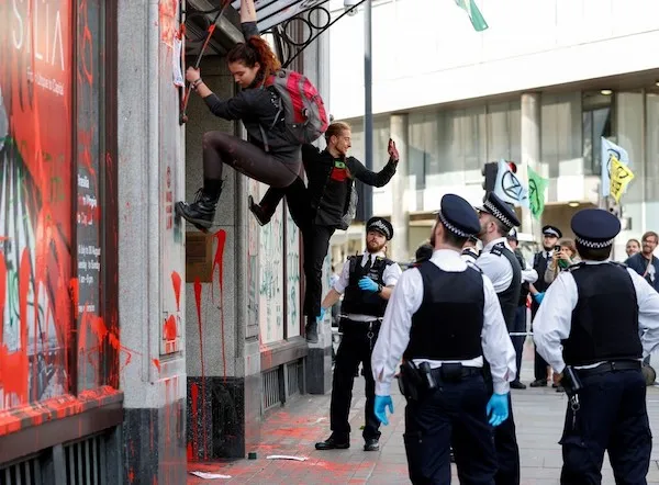 Osoby protestujúce pri budove s nápisom, policajti sledujú. Protestné akcie s transparentom na pozadí.