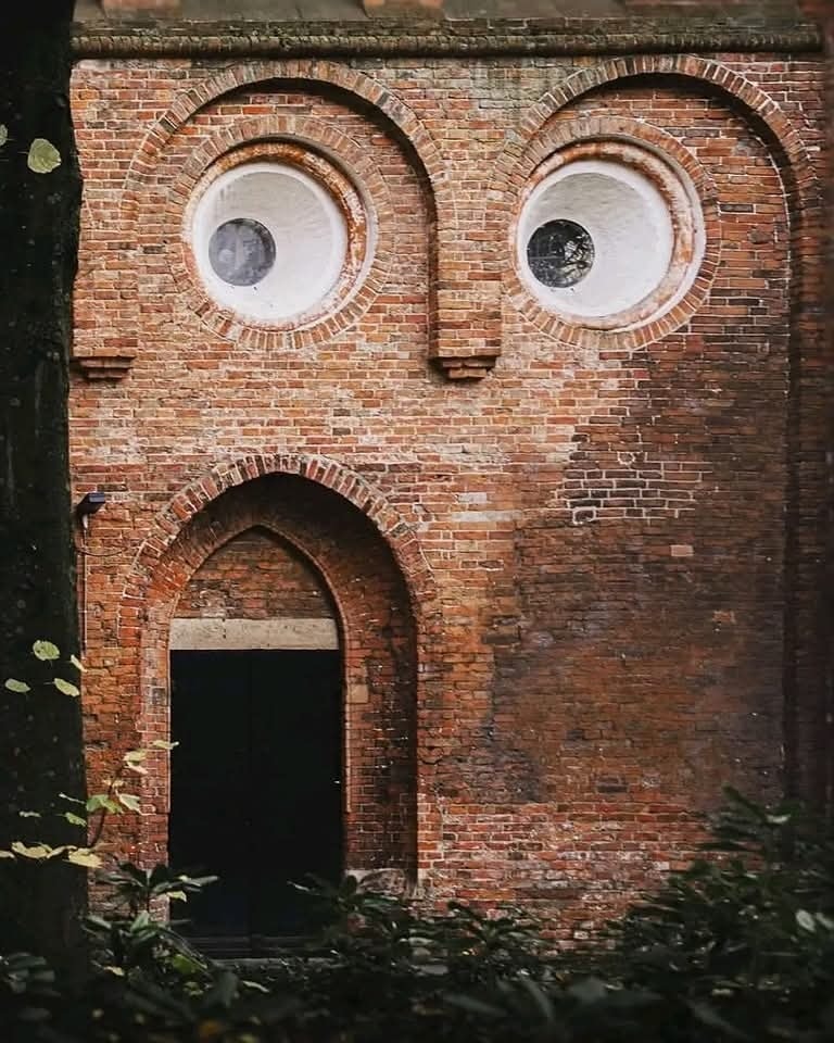 Stará tehlová budova s oknami a dverami, ktorá pripomína ľudskú tvár, obklopená prírodou.