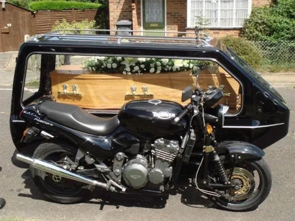 Motorka s prívesom na rakvu zdobenou kvetmi, zaparkovaná pred budovou na pokojnej ulici.