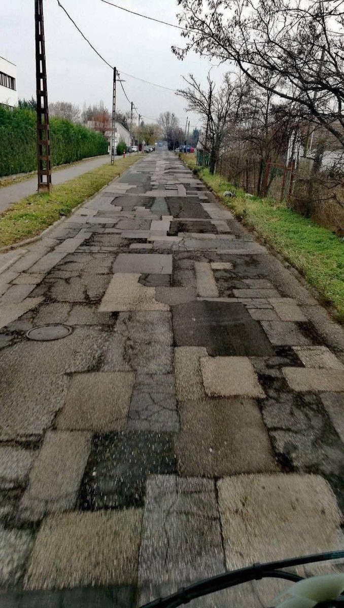 Nerovná asfaltová cesta s mnohými záplatami vedie popri stromoch a budovách v jesennej krajine.