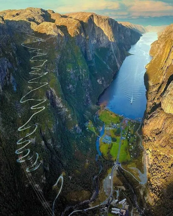 Zákrutová cesta na útesoch nad fjordom s plávajúcou loďou, obklopená strmými skalami a malebným údolím.