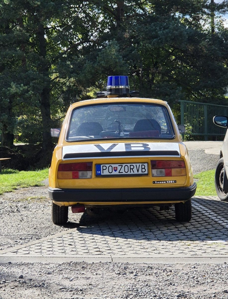Žlté vozidlo Škoda 120L s nápisom VB a modrým majákom zaparkované na slnečnom dvore medzi stromami.