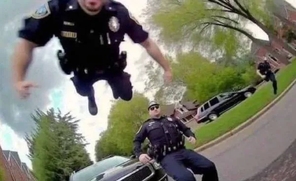 Policajt v skoku nad autom, ďalší policajti stoja na ceste pri zeleni a domoch.