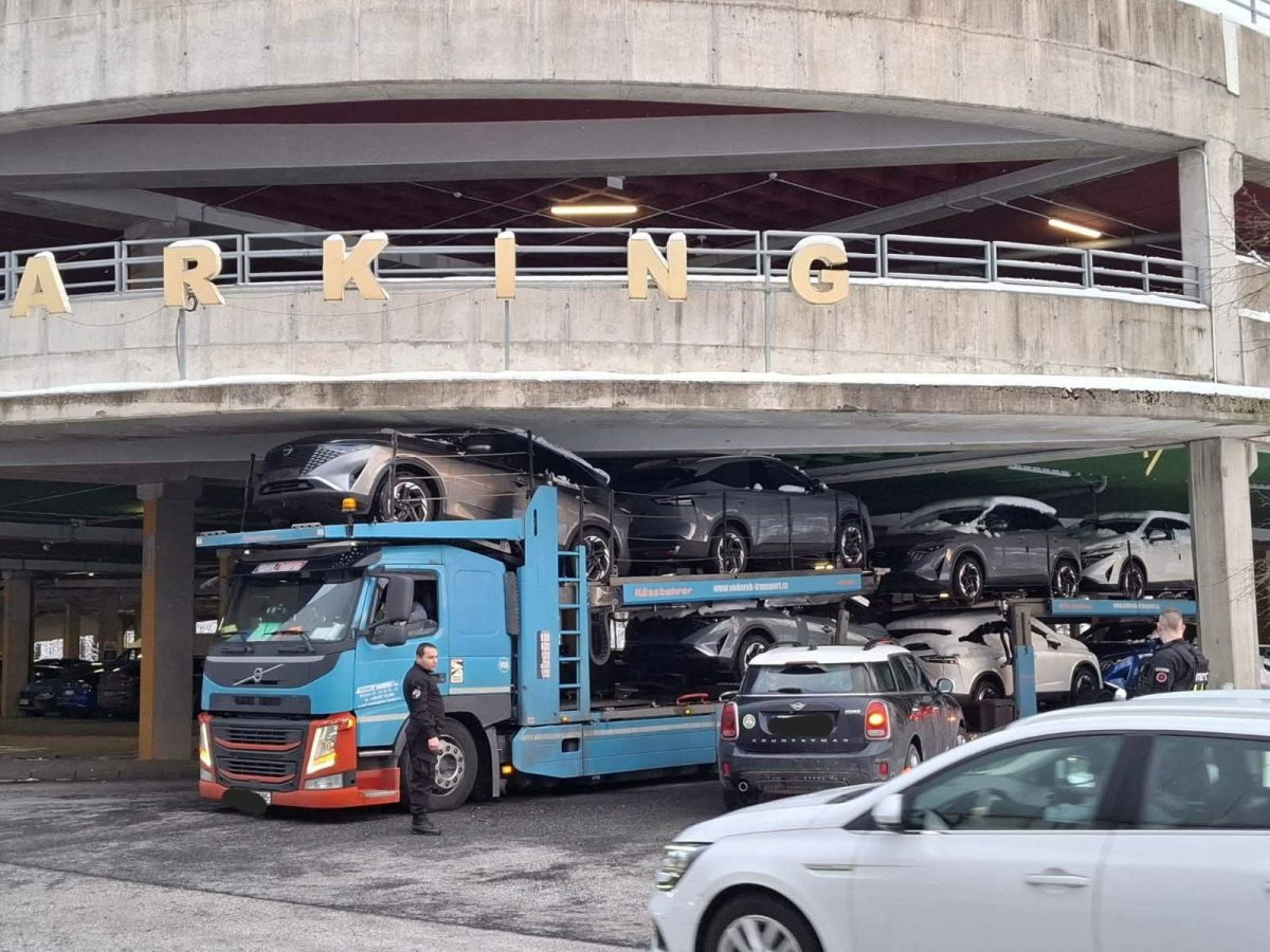 Kamión prevážajúci viacero automobilov zastavený pred parkovacou garážou, zatiaľ čo okolo prechádzajú autá.