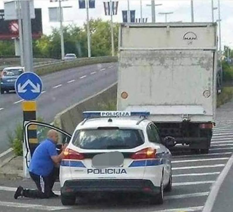 Policajné auto stojí za kamiónom na ceste, policajt kľačí pri aute a kontroluje situáciu.