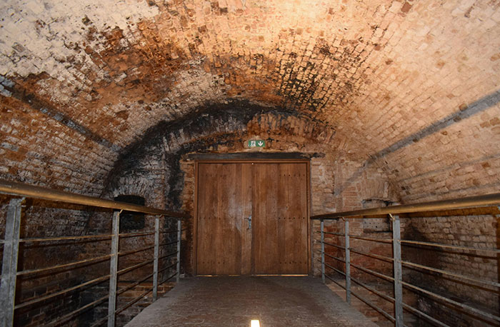 Starobylý tehlový klenutý tunel s drevenými dverami a kovovým zábradlím, osvetlený jemným vnútorným svetlom.