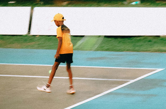 Chlapec v žltom hrá tenis na ihrisku, sústredený na hru. Farby a pohyb vytvárajú dynamickú atmosféru zápasu.
