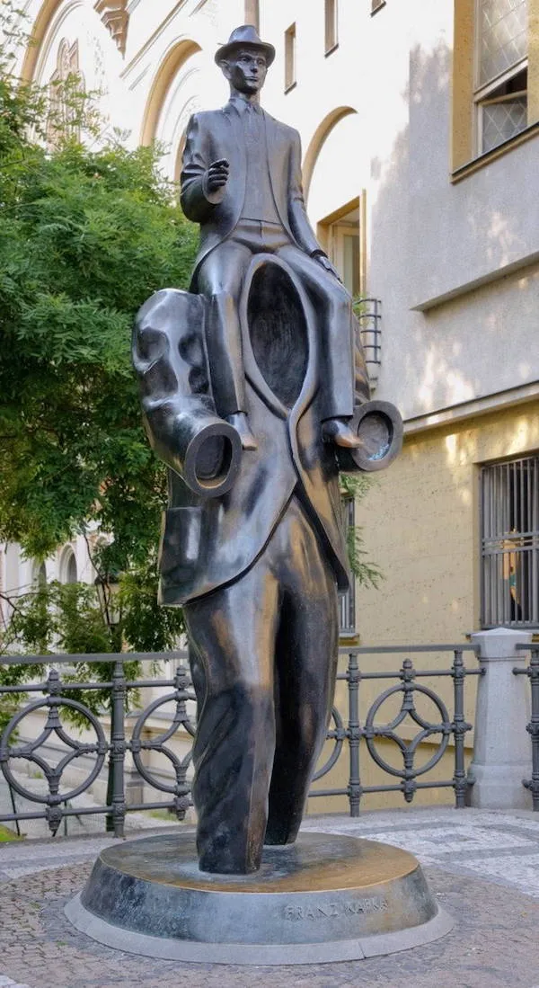 Socha muža sediaceho na obrovskej nohavici bez hlavy, umiestnená v mestskom parku s budovou v pozadí.