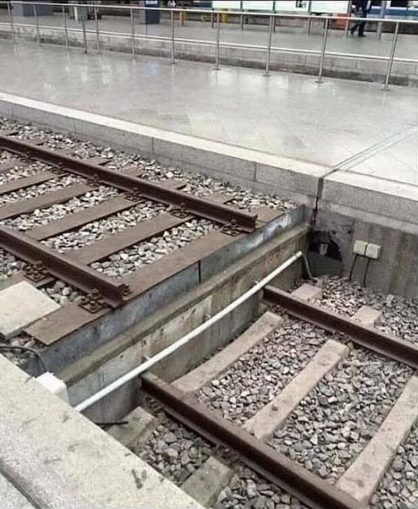 Nedostavaná železničná trať s chýbajúcim mostom, vedúca na perón v stanici, ilustruje nedokončenú infraštruktúru.