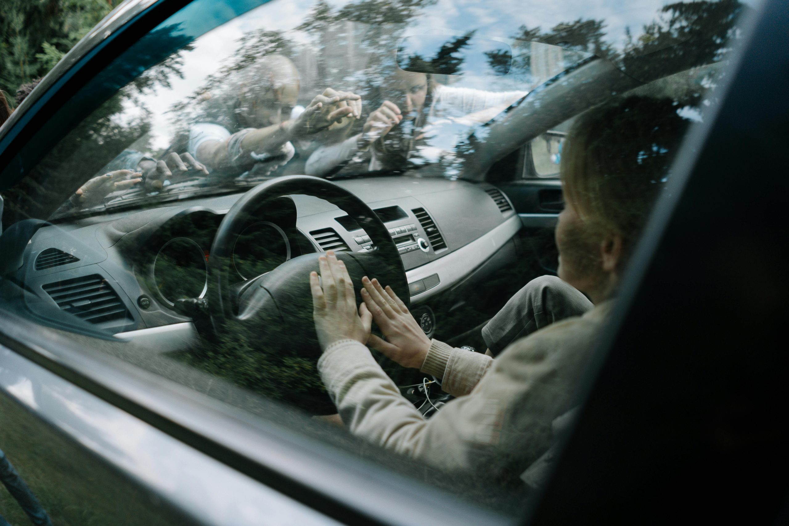 Osoba zatvorená v aute, stlačené ruky na volante, ľudia na kapote vonku, napätá situácia.