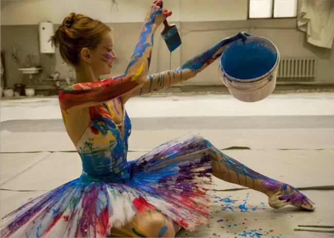 Baletka pokrytá farebnou farbou sedí na zemi a prevracia modrú farbu z vedra, umelecký performance.