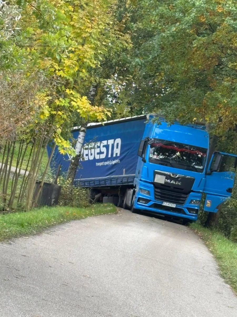 Nákladné auto s modrým návesom uviazlo vo svahu úzkej vidieckej cesty obklopenej stromami.