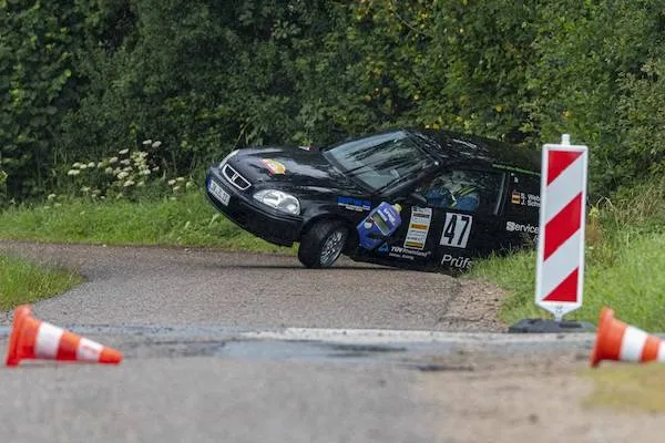 Auto číslo 47 uviaznuté v príkope na rally preteky, obklopené kužeľmi na ceste, hustý les v pozadí.