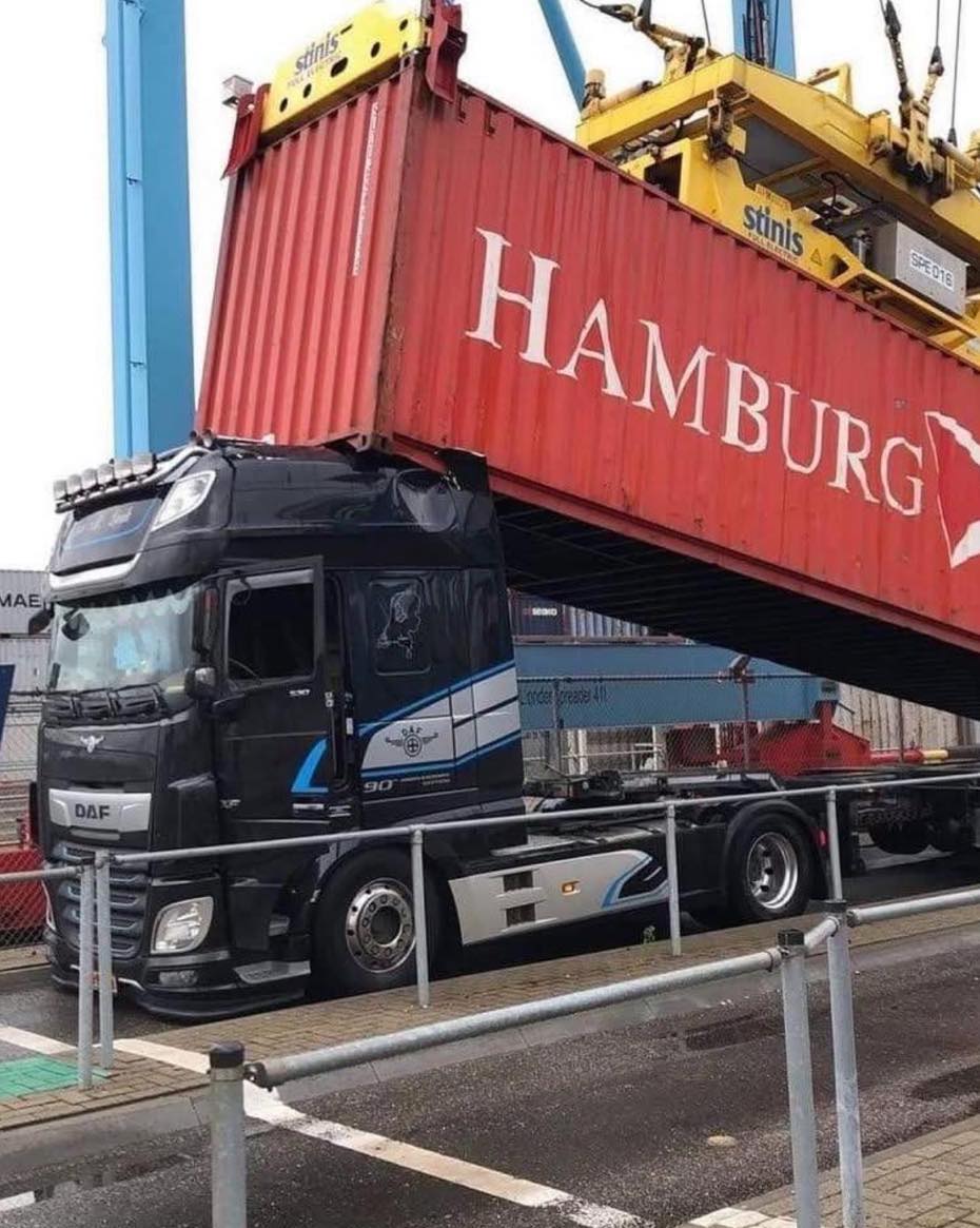 Nákladné auto uviaznuté pod kontajnerom v prístave, nápis Hamburg na červenom kontajneri s modrým žeriavom.