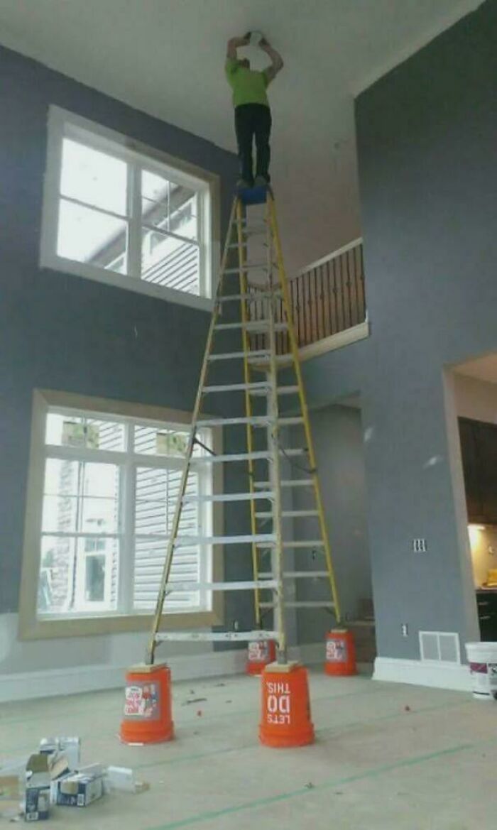 Osoba na vysokej rebríku v interiéri stojí na vedrách, vykonáva údržbu stropu.