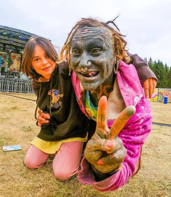 Dieťa a osoba s výraznými tetovaniami pózujú na hudobnom festivale, s priateľským gestom pokoja.
