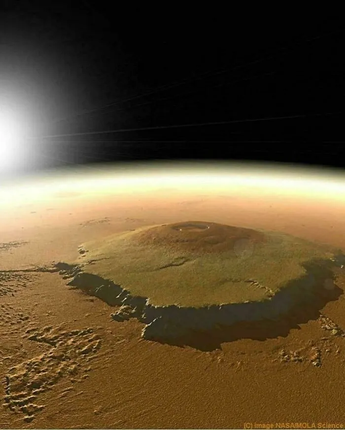 Povrch Marsu s pohľadom na veľkú sopku a horizont v diaľke, slnečný svit zľava.