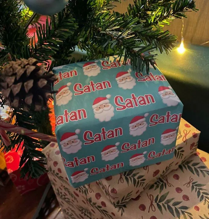 Darček pod stromčekom zabalený v modrom papieri s nápisom Satan namiesto Santa.