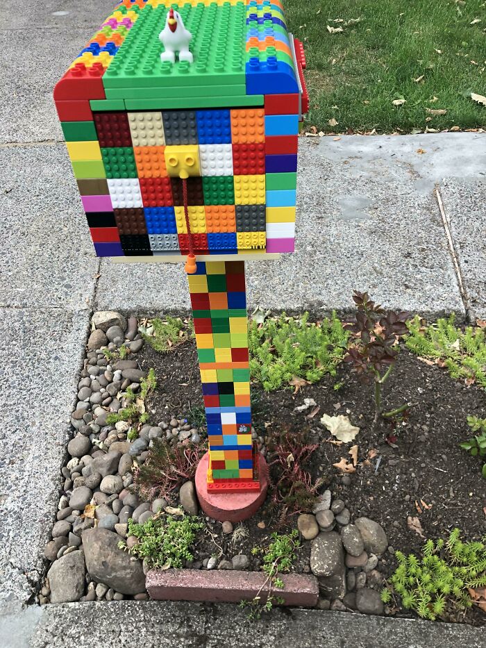 Poštová schránka z LEGO kociek na záhrade, zdobená malou plastikovou kurou na vrchu.