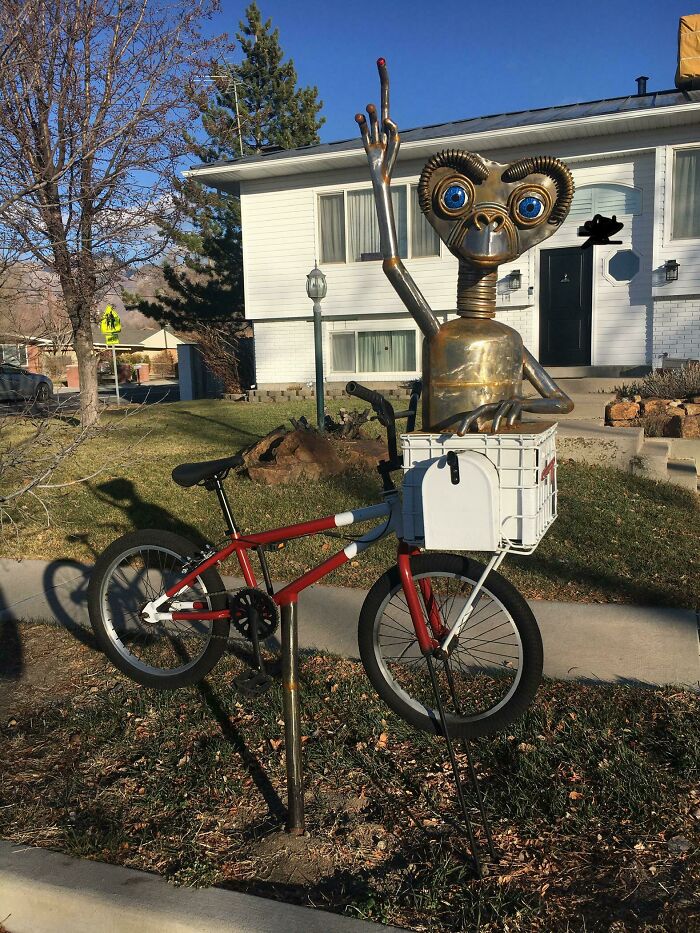 Socha mimozemšťana s bicyklom pred domom počas slnečného dňa, originálna vonkajšia dekorácia.