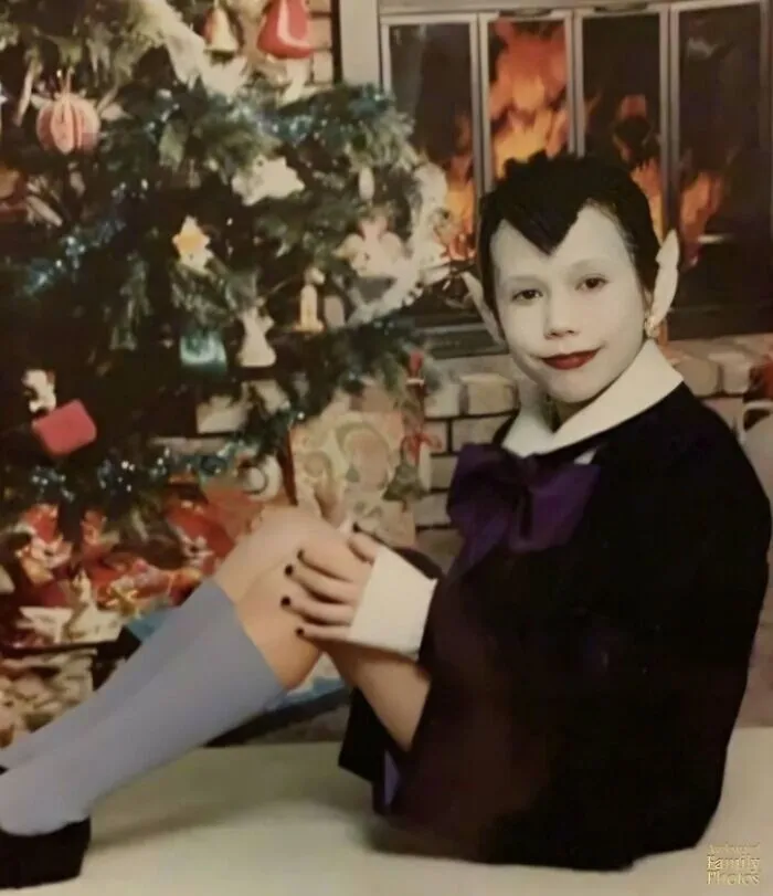 Dieťa v kostýme upíra sedí pri vianočnom strome, s plameňmi v kozube v pozadí.