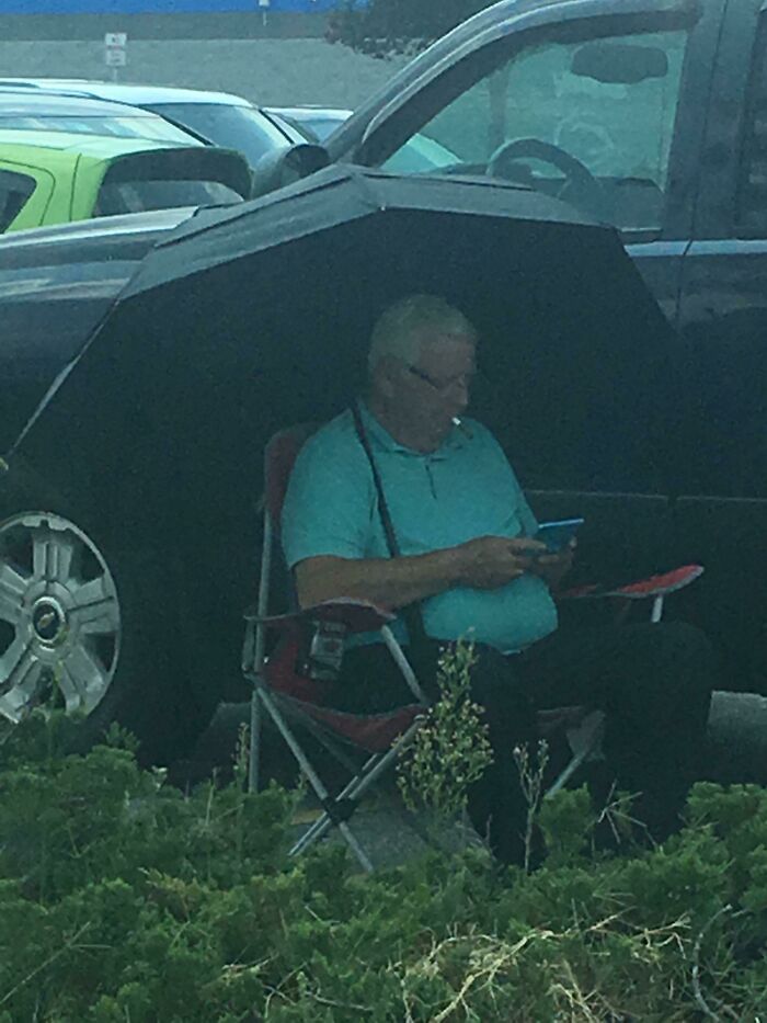 Neznámy muž sedí na stoličke vonku pod dáždnikom a používa svoj mobilný telefón na parkovisku.