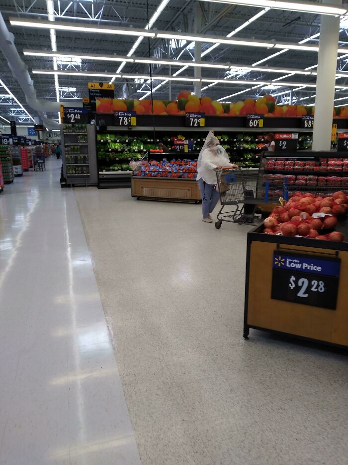 Osoba v supermarkete tlačí vozík s potravinami, oblečená v plastovej ochrane, prechádzajúc okolo výstav s ovocím.