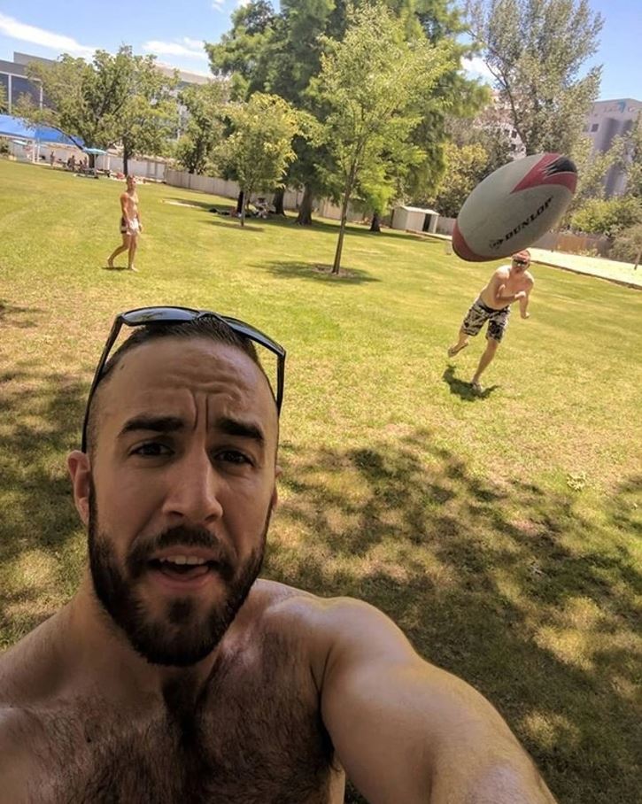 Muž robí selfie na trávniku, zatiaľ čo iný muž chytá loptu v parku za slnečného dňa.