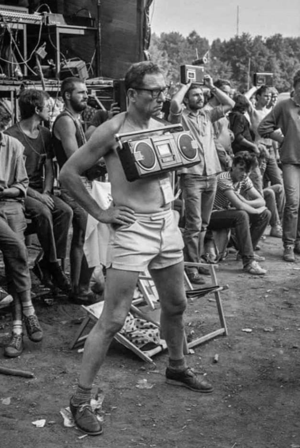 Muž s rádiom na hrudi stojí v skupine ľudí vonku, oblečený v krátkych nohaviciach. Retro festivalová atmosféra.