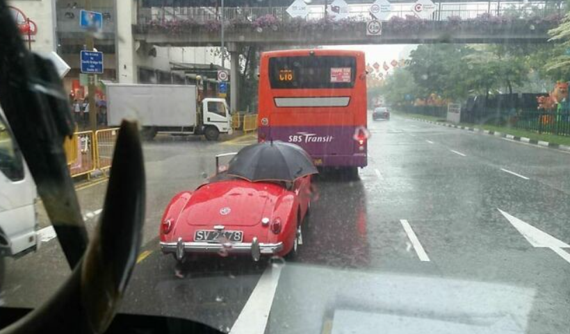 Červené retro auto s otvoreným dáždnikom, stojace na dažďovej ceste za autobusom.