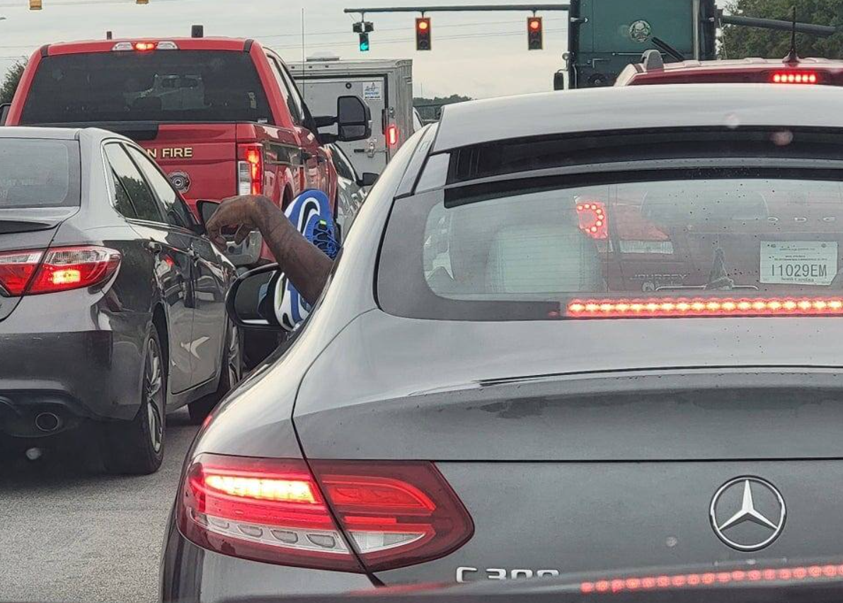 Orezaná ruka s umelou dinosaurou figúrkou smeruje z okna auta na rušnej križovatke medzi autami Mercedes a Ford.