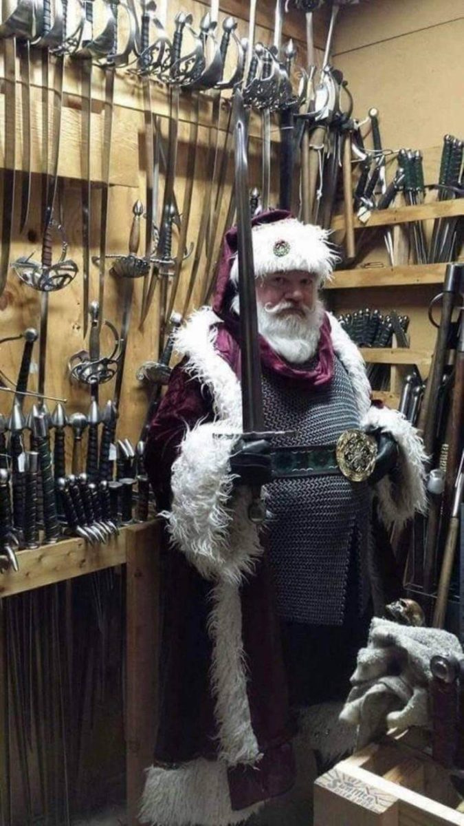 Muž v kostýme s vianočnou čiapkou drží meč v miestnosti plnej zbraní.