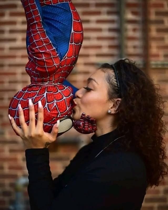 Osoba pobozkáva dieťa v kostýme Spidermana, visí dolu hlavou s tehlovou stenou v pozadí.