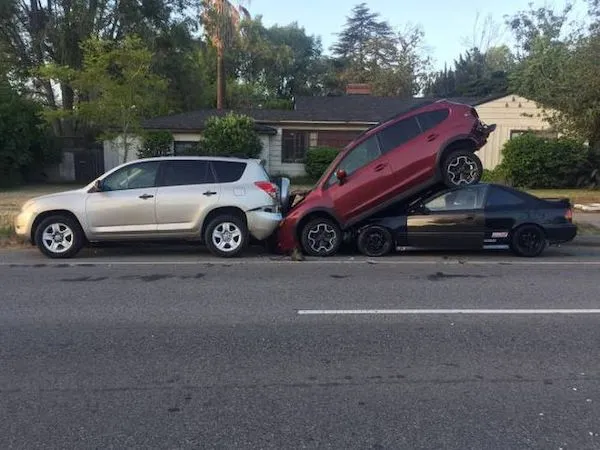 Tri autá v reťazovej dopravnej nehode, pričom červené vozidlo je naklonené na čierne auto na mestskom okraji.