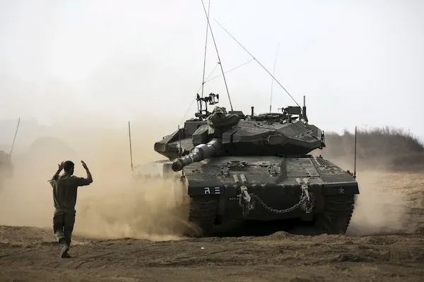 Vojenský tank v pohybe na piesočnej pôde s vojakom usmerňujúcim jeho trasu.