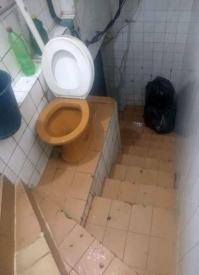 Malá kúpeľňa s toaletou na úrovni schodov a keramickými dlaždicami, vedľa plastových fliaš a smetného koša.