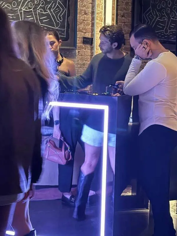 Muži a ženy pri pódiu v klube s moderným osvetlením a neformálne oblečenými návštevníkmi.