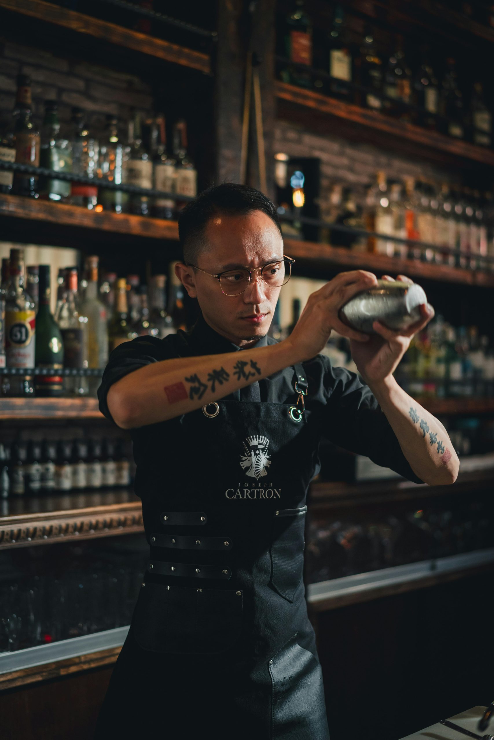 Barman mieša koktail v štýlovom prostredí, obklopený rôznymi druhmi alkoholu na poličkách.
