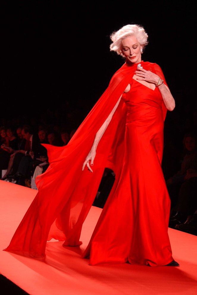 Elegantná staršia modelka v červenej róbe kráča na móle počas módnej prehliadky.