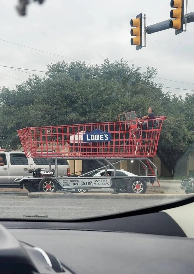 Obrovský nákupný vozík s logom Lowe's na podvozku auta na rušnej ceste na križovatke.