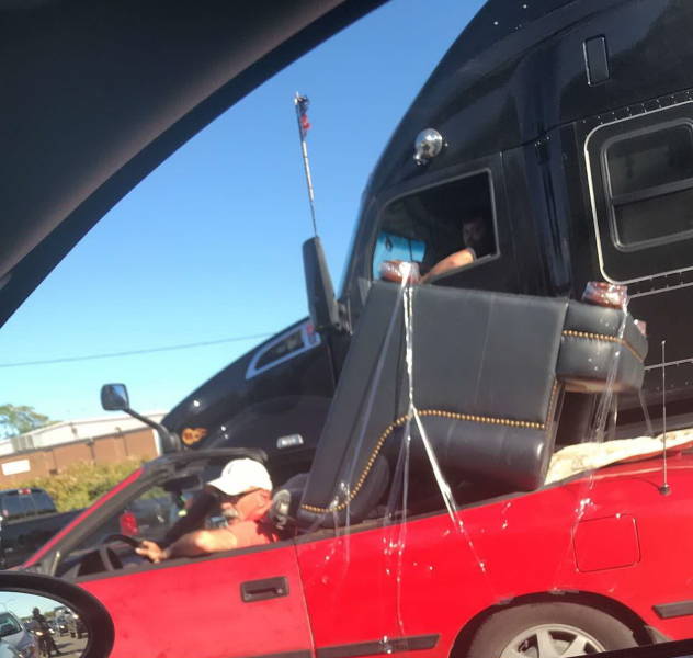 Auto nesie veľkú čiernu sedačku na ceste, pričom vodič vedľa v nákladnom aute pozoruje situáciu.