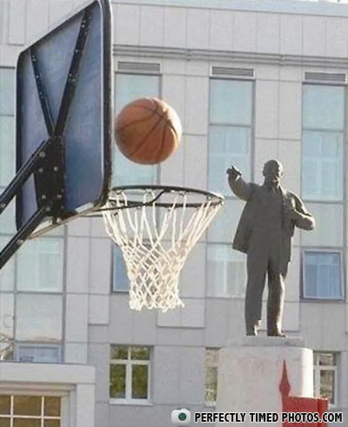 Socha ukazuje na basketbalovú loptu letiacu do koša pred budovou.