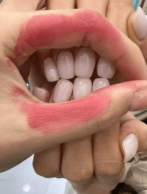 Ruky tvarujúce ústa s rúžom okolo, pričom v strede sú vidieť biele nechty, tvorivé nechtové umenie.