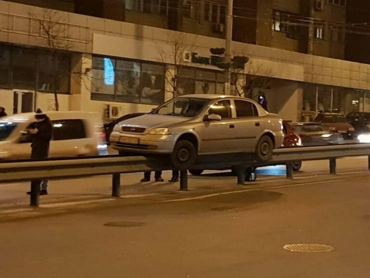 Auto zaparkované na zvodidle na večernej ulici, okoloidúci pozorujú situáciu.