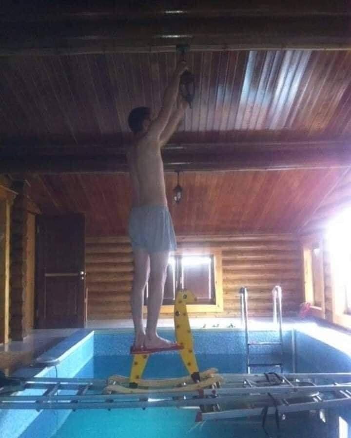Muž na neistom rebríku v interiéri bazéna robí domáce práce pod dreveným stropom.