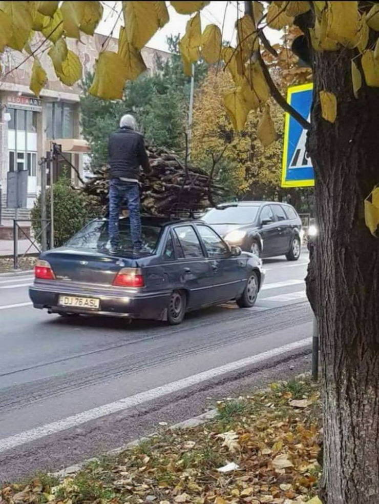 Osoba stojaca na aute prepravuje veľkú kopu dreva cez jesennú ulicu.