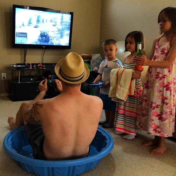 Muž sedí v detskom bazéniku, hrá videohru s deťmi okolo, zatiaľ čo na TV beží hra.