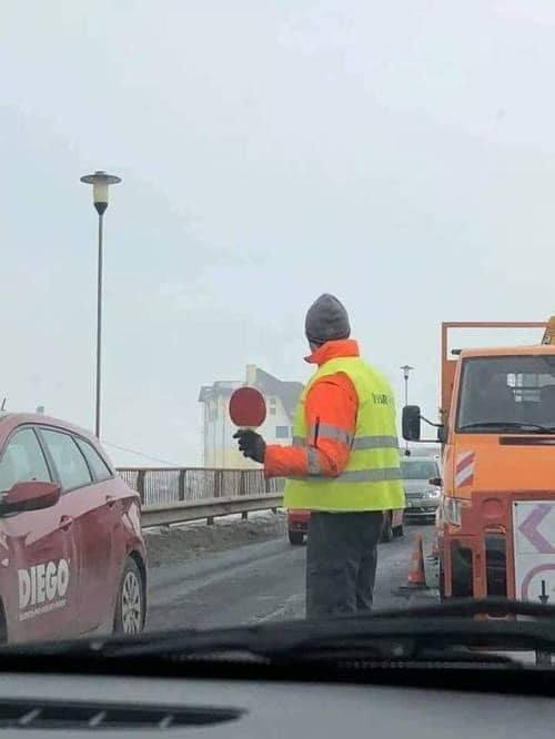 Pracovník v reflexnej veste riadi dopravu na ceste vedľa oranžového nákladného auta.