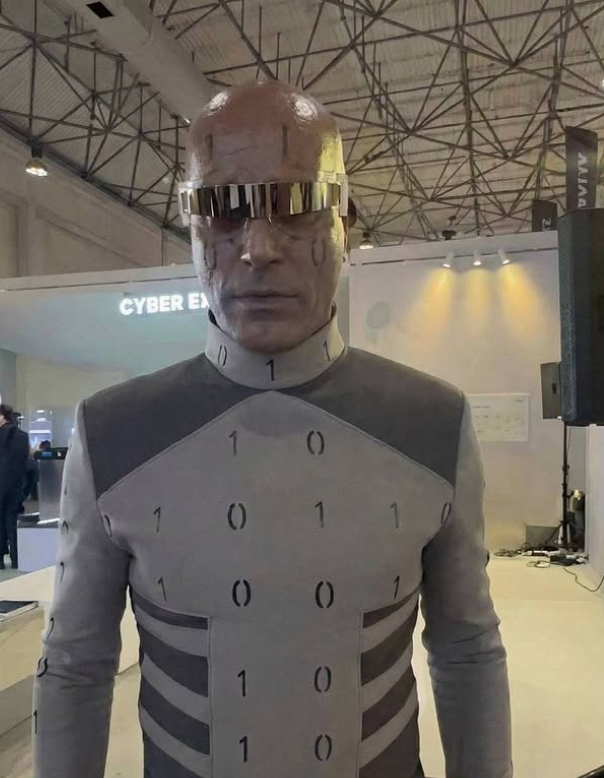 Ľudská postava v futuristickom obleku s binárnym vzorom a zrkadlovými okuliarmi na technologickom podujatí.